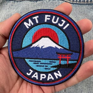 May include: A circular embroidered patch featuring a dark blue border with the white text MT FUJI and JAPAN. The center depicts a snow-capped mountain against a red sun and light blue sky, with a red torii gate standing in blue water.