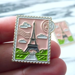 May include: A pink and gold enamel pin in the shape of a postage stamp, depicting the Eiffel Tower, clouds, and a crescent moon. The pin has a gold border and a white road.