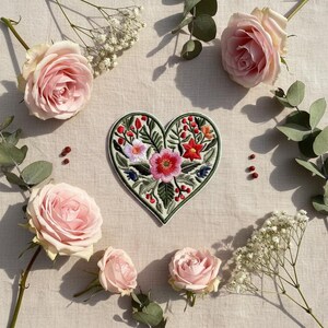 Può includere: Una toppa ricamata a forma di cuore con un disegno floreale, con fiori rosa, rossi e blu, circondati da foglie verdi. La toppa è ambientata tra rose rosa, rametti di gipsofila e foglie di eucalipto su uno sfondo neutro.