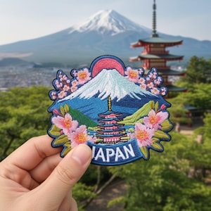 Peut inclure: Écusson brodé représentant le Mont Fuji avec un sommet enneigé, un soleil rouge et une pagode à plusieurs niveaux. Des fleurs de cerisier roses et le mot "JAPAN" en blanc complètent le motif. L'écusson est tenu devant un arrière-plan panoramique.