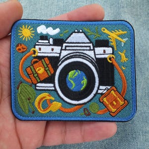 May include: Embroidered patch depicting a classic camera with a globe in the lens. The design incorporates a sun, aeroplane, and luggage in bright colours. The patch has a blue border and is held in a hand.