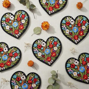 Pode incluir: Patches bordados em forma de coração com borda preta, apresentando um design floral colorido em vermelho, azul, branco e verde. Os patches são dispostos em uma superfície de cor clara, com flores secas e vegetação espalhadas.