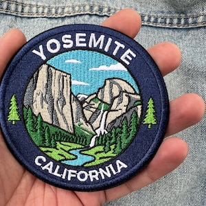 May include: A circular embroidered patch featuring a landscape of Yosemite, California. The design includes granite cliffs, a waterfall, a blue river, and green pine trees against a light blue sky. The text YOSEMITE and CALIFORNIA is in white.