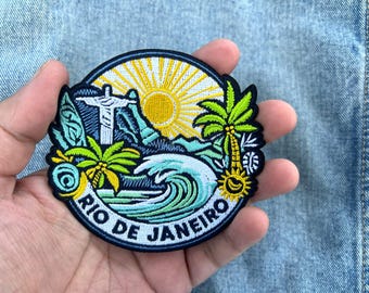 May include: Embroidered patch featuring a Rio de Janeiro design. The patch has a circular shape with a blue border and depicts the Christ the Redeemer statue, palm trees, a sun, and ocean waves. The text "RIO DE JANEIRO" is also included.