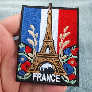 May include: Embroidered patch depicting the Eiffel Tower in gold, set against the French flag's blue, white, and red. The word "FRANCE" is at the base, with floral details in red, blue, and beige.