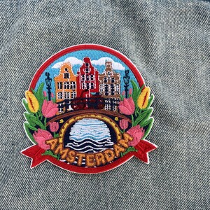 Puede incluir: Parche bordado con una colorida escena de &Aacute;msterdam. El dise&ntilde;o incluye casas ic&oacute;nicas de los canales, un puente y tulipanes, con la palabra "AMSTERDAM" en una pancarta roja. El parche est&aacute; sobre un fondo de mezclilla.