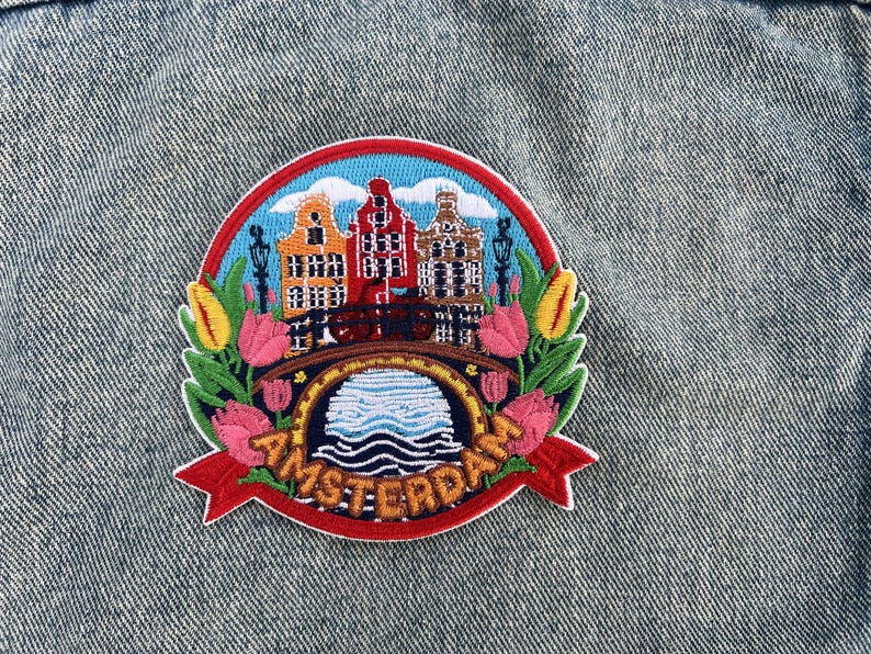 Puede incluir: Parche bordado con una colorida escena de &Aacute;msterdam. El dise&ntilde;o incluye casas ic&oacute;nicas de los canales, un puente y tulipanes, con la palabra "AMSTERDAM" en una pancarta roja. El parche est&aacute; sobre un fondo de mezclilla.