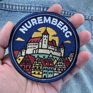 May include: A circular embroidered patch featuring a detailed depiction of the Nuremberg castle and city skyline. The design includes red and yellow buildings against a dark blue background with the word NUREMBERG stitched in white at the top.