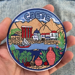 May include: Circular embroidered patch depicting Crete, with the word "CRETE" in blue. The design shows mountains, buildings, a sailboat, and pottery in red, blue, and beige, with a white border. The patch is held in a hand.