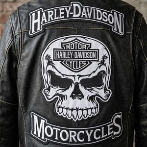 Puede incluir: Chaqueta de moto de cuero negro con parches Harley-Davidson. La parte trasera presenta un gran parche de calavera blanca con las palabras "Harley-Davidson Motorcycles" y el logotipo de Harley-Davidson.