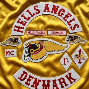 Puede incluir: Una colección de parches bordados sobre una tela de satén amarilla. Los parches incluyen un texto arqueado grande 'HELLS ANGELS', un rocker 'DENMARK', un emblema central de calavera alada, parches de diamante 'MC' y '1%', e insignias de calavera alada más pequeñas.