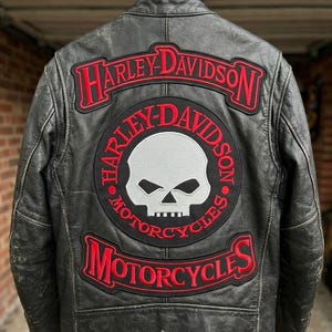 Può includere: Giacca da moto in pelle nera con toppe Harley-Davidson. La giacca presenta loghi Harley-Davidson rossi e bianchi e una grafica a teschio sul retro. La giacca è appesa a una gruccia.