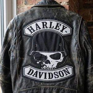 Puede incluir: Chaqueta de cuero negro desgastado con un gran parche de Harley-Davidson en la espalda. El parche presenta una calavera con casco, con el nombre de la marca en letras blancas. La chaqueta está colgada de una percha metálica.