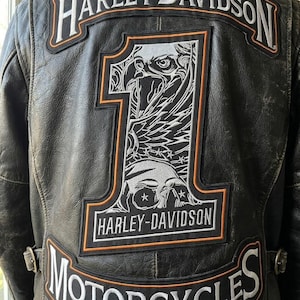 以下が含まれることがあります： ハーレーダビッドソンのパッチが付いた黒い革のバイクジャケット。背面には、ワシのデザインと数字の1が入った大きなパッチ、ハーレーダビッドソンとモーターサイクルのロゴが白とオレンジ色で表示されています。