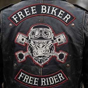 Op de afbeelding: Zwarte leren jas met een groot geborduurd embleem. Het embleem toont een gorilla met een bril en gekruiste zuigers. De tekst "FREE BIKER" staat erboven en "FREE RIDER" eronder in wit met rode rand.