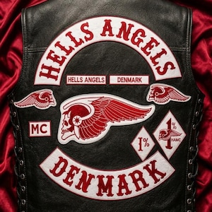Puede incluir: Un chaleco de cuero negro con parches bordados en blanco y rojo. La parte posterior muestra el texto HELLS ANGELS y DENMARK en grandes pancartas arqueadas, con un emblema central de calavera alada, parches de calaveras, MC y diamantes 1%.