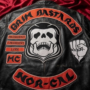 Puede incluir: Una chaqueta de cuero negro con parches bordados. La parte posterior muestra una calavera grande, el texto GRIM BASTARDS, NOR-CAL, PRESIDENT, V.PRESIDENT, LODI y MC en naranja y negro, junto a un parche de un puño blanco a la derecha.