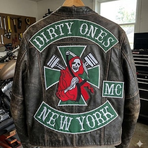 Puede incluir: Chaqueta de cuero marrón con parches. La parte trasera presenta las palabras "DIRTY ONES NEW YORK MC" en blanco sobre verde, y un diseño de la Parca en rojo y blanco.