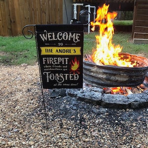 May include: A black and yellow welcome sign with the text "Welcome to The Anore's Firepit where friends and marshmallows get toasted at the same time!" hanging on a metal stake in front of a fire pit with flames.