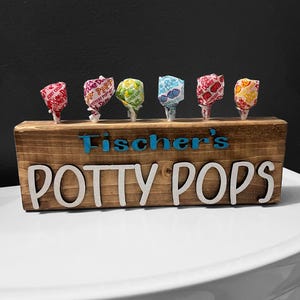 May include: A wooden display with the text "Fischer's POTTY POPS" in white and blue lettering. Six wrapped lollipops in various colors are inserted into the wooden block. The lollipops have different colored wrappers.
