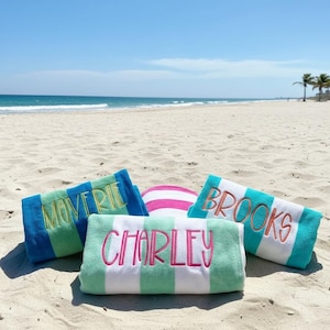 May include: Beach towels with personalized names embroidered in various colors. The towels are striped in blue, green, and white, with names like "MOVERIE", "BROOKS", and "CHARLEY" in contrasting colors. A pink and white striped beach ball is also present.