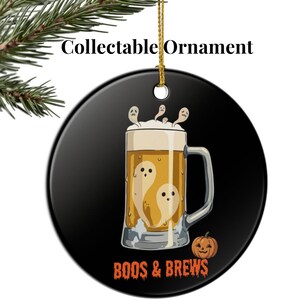 Può includere: Un ornamento rotondo in ceramica nera con la grafica di un boccale di birra pieno di liquido dorato e piccoli fantasmi bianchi. Il testo "BOOS & BREWS" appare in arancione in basso, con altri fantasmi sopra la schiuma e una piccola zucca.