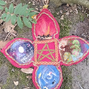 Op de afbeelding: Een kruisvormige vilten wandhanger met een centraal vierkant met een pentagram, omringd door vier bloembladachtige secties die een kampvuur, een nachtelijke hemel met een maan, een groene boom en een blauwe waterscène met visjes uitbeelden.
