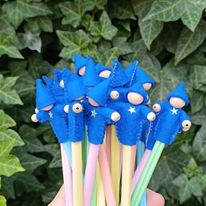Peut inclure: Un bouquet de crayons surmontés de figurines de sorciers en feutrine bleue. Chaque personnage possède un chapeau pointu bleu, une tête en perle de bois et une étoile blanche appliquée sur la poitrine. Les crayons ont des corps pastel rose, jaune et bleu.