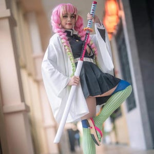 May include: A cosplay outfit featuring a white haori, a dark uniform with a pleated skirt, and green striped socks. The ensemble includes a long, pink and lime green braided wig and a long, thin sword with a pink blade and a blue-wrapped handle.