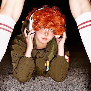 May include: An olive green hooded jacket with a front zipper, worn with white knee-high socks featuring two red stripes. The outfit includes white over-ear headphones and a short, curly, vibrant orange wig, complemented by light freckles on the face.