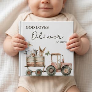 May include: A personalized children's book with the text "GOD LOVES Oliver SO MUCH" on the cover. The cover features a watercolor illustration of a tractor pulling a wagon with farm animals. The book is held by a baby.