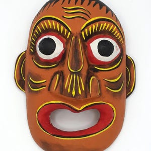 Puede incluir: Una máscara marrón pintada a mano con detalles en negro, amarillo y rojo. La máscara presenta ojos grandes y negros con contornos blancos, una boca roja ancha y rasgos faciales estilizados. El diseño incluye líneas y detalles amarillos.