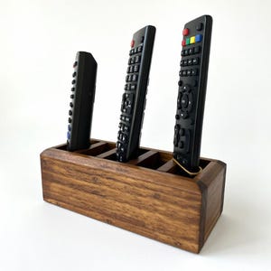 Könnte beinhalten: Ein Fernbedienungsorganizer aus Holz mit vier Fächern, der drei schwarze Fernbedienungen aufnimmt. Der Organizer ist aus gebeiztem Holz und hat eine rechteckige Form. Ein Gummiband sichert eine Fernbedienung.