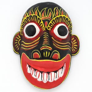 May include: A hand-painted wooden mask with a black base, featuring large white-rimmed eyes and a wide red mouth with white teeth. Yellow and red accents highlight the eyebrows, ears, and facial details. The mask has a smiling expression.