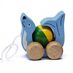 May include: A blue wooden pull toy shaped like a seal, holding a colorful ball. The toy has two wooden wheels and a white string for pulling. The ball is yellow, green, and red.