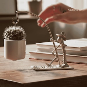 May include: A decorative metal sculpture of a person fishing for a cactus in a white pot. The sculpture is on a wooden table with books in the background. The text "PLANT LOVER GIFT" is visible.