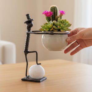 May include: A dark metal sculpture of a figure wearing a top hat, standing on a square base with one foot on a white stone. The figure holds a white ceramic bowl containing a green cactus with pink flowers and a green succulent plant.