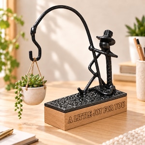 Könnte beinhalten: Eine schwarze Metallskulptur einer Figur mit Hut, die auf einem rechteckigen Holzsockel mit der Aufschrift A LITTLE JOY FOR YOU sitzt. Die Figur hält eine gebogene Stange mit einem Haken, an dem ein kleiner weißer Keramiktopf mit Sukkulente hängt.