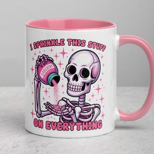Könnte beinhalten: Weiße Keramiktasse mit rosa Innenraum und Henkel. Die Tasse zeigt ein Cartoon-Skelett, das rosa Pulver aus einem Behälter mit der Aufschrift "Sarcasm" gießt. Der Text auf der Tasse lautet "I sprinkle this stuff on everything."