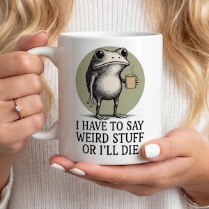 Op de afbeelding: Witte keramische mok met een grillige illustratie van een kikker die een koffiekopje vasthoudt. De kikker staat tegen een groene cirkel, met daaronder de tekst "I HAVE TO SAY WEIRD STUFF OR I'LL DIE". Een leuk, eigenzinnig ontwerp.