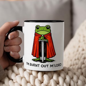Può includere: Una tazza in ceramica bianca con manico e bordo neri, raffigurante una rana verde che indossa un mantello rosso e impugna una spada argentata. Sotto la rana, il testo "I'M BURNT OUT M'LORD" è scritto in lettere maiuscole nere.