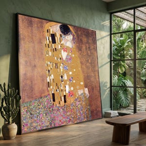 Puede incluir: Gran impresión enmarcada de "El Beso" de Gustav Klimt. La obra representa a una pareja abrazándose, con figuras adornadas con oro y patrones coloridos. El fondo es marrón texturizado, y el marco es negro.