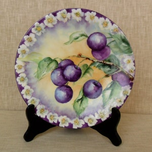 May include: A vintage porcelain plate with a hand-painted design of purple plums on a branch with green leaves. The plate has a white and purple border with a floral pattern.