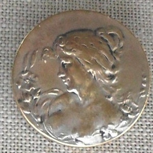 May include: A silver and gold toned metal button with a detailed design of a woman's profile with flowing hair and a floral motif.