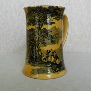 May include: A yellow ceramic mug with a black and white landscape scene and figures. The mug has a handle and a wide base.