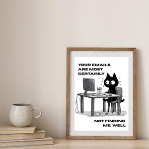 May include: A framed print featuring a black and white cartoon of a cat at a desk, looking anxious. The text on the print says "YOUR EMAILS ARE MOST CERTAINLY NOT FINDING ME WELL." A mug and books are on the table.