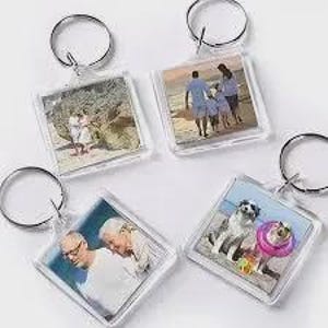 May include: Four square clear acrylic keychains, each featuring a different photograph insert. The images include a couple on a beach, a family walking on the sand, two older individuals, and a dog wearing a pink floatie on a beach.