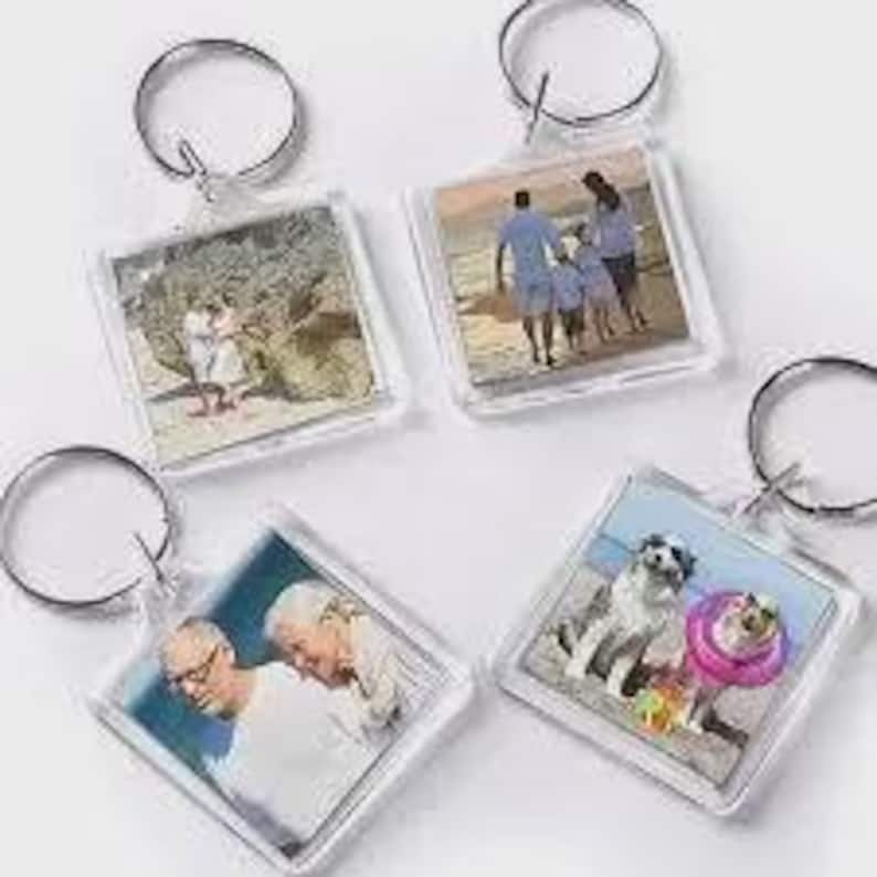 May include: Four square clear acrylic keychains, each featuring a different photograph insert. The images include a couple on a beach, a family walking on the sand, two older individuals, and a dog wearing a pink floatie on a beach.