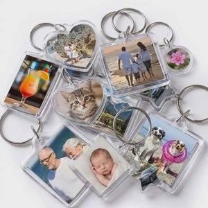 May include: A collection of clear acrylic keychains in various shapes including hearts, squares, and stars, each containing a different photograph. The images feature people on a beach, a kitten, a cocktail, a baby, a dog, and a pink flower.