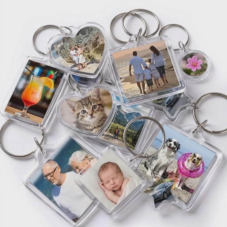 May include: A collection of clear acrylic keychains in various shapes including hearts, squares, and stars, each containing a different photograph. The images feature people on a beach, a kitten, a cocktail, a baby, a dog, and a pink flower.
